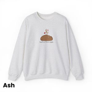 Peut inclure: Un sweat-shirt gris clair &agrave; col rond. Il pr&eacute;sente une illustration de pain avec de la vapeur et deux c&oelig;urs, ainsi que le texte "loaf at first sight" en dessous. Le mot "Ash" est en bas.