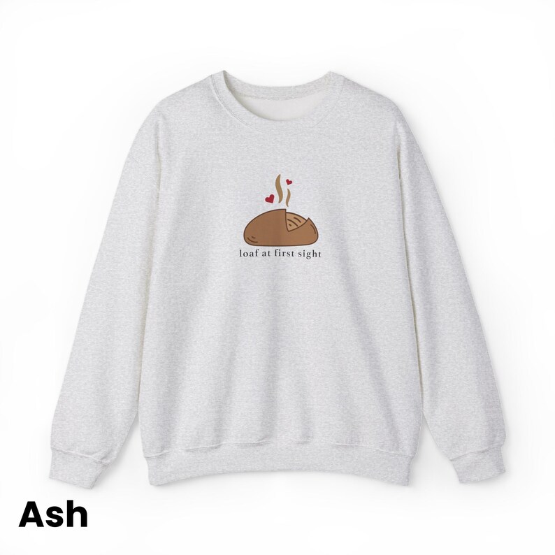 Peut inclure: Un sweat-shirt gris clair &agrave; col rond. Il pr&eacute;sente une illustration de pain avec de la vapeur et deux c&oelig;urs, ainsi que le texte "loaf at first sight" en dessous. Le mot "Ash" est en bas.