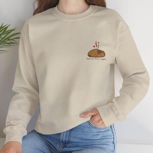 May include: Beige sweatshirt with a crew neck. The design features a cartoon loaf of bread with steam and hearts, and the text "loaf at first sight" in a small font below the bread. The sweatshirt is a neutral color.