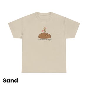 Puede incluir: Camiseta color arena con un gr&aacute;fico de una hogaza de pan con vapor y corazones, y el texto "loaf at first sight". Es una prenda de vestir informal.