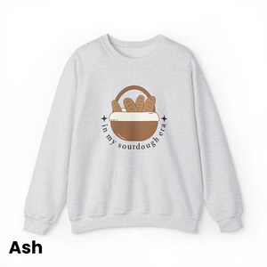 Peut inclure: Sweat-shirt gris cendr&eacute; &agrave; col rond. L'avant pr&eacute;sente un motif de panier marron rempli de pains, avec le texte "in my sourdough era" dans un design circulaire. Le mot "Ash" est imprim&eacute; en bas.