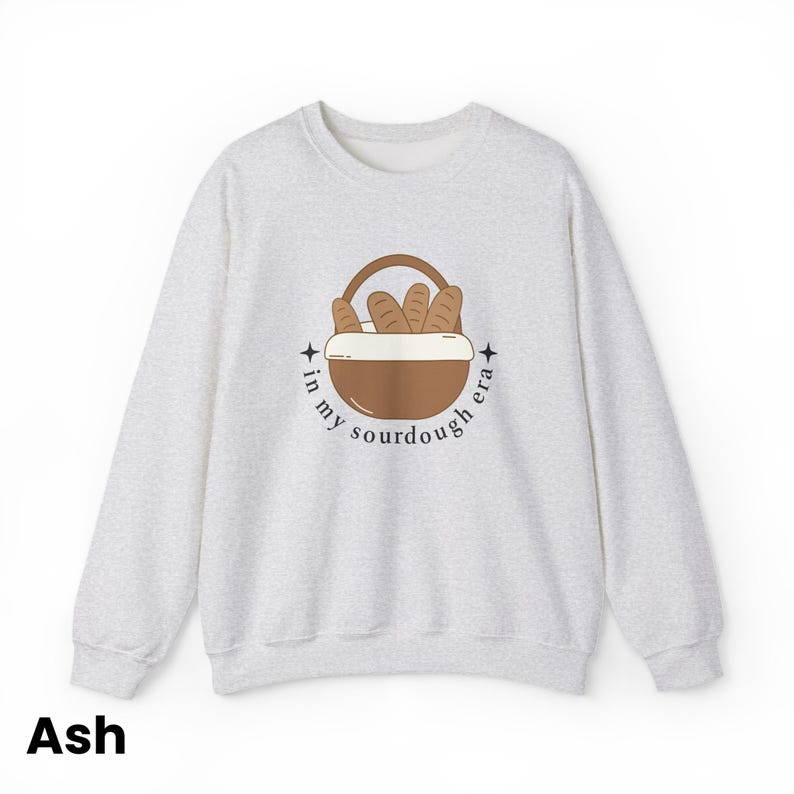 Peut inclure: Sweat-shirt gris cendr&eacute; &agrave; col rond. L'avant pr&eacute;sente un motif de panier marron rempli de pains, avec le texte "in my sourdough era" dans un design circulaire. Le mot "Ash" est imprim&eacute; en bas.