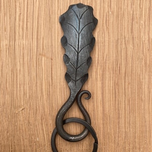 Puede incluir: Llavero de metal negro con un diseño de hoja de roble. La hoja está detallada con venas y tiene un tallo rizado. El llavero tiene un anillo para sujetar las llaves.