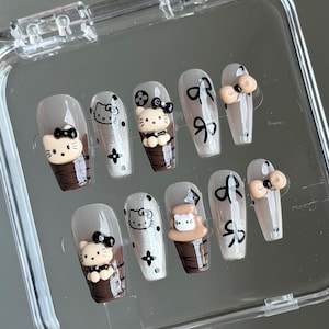 May include: A set of ten artificial nails with a clear, glossy finish. The nails feature various designs, including a Hello Kitty character, bows, and a brown cup design. The nails are in a clear plastic case.