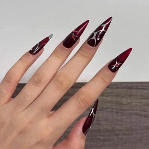 May include: A hand displaying long, stiletto-shaped artificial nails. The nails are a deep burgundy color with silver and red accents. Some nails feature silver swirls and red gem-like embellishments, creating a striking and elegant design.
