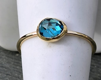 May include: A gold ring featuring a single, round, faceted blue gemstone set in a smooth, polished gold bezel. The band is thin and circular, showcasing a minimalist design that highlights the deep blue color and reflective surface of the stone.
