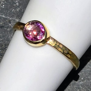 Op de afbeelding: Een gouden ring met een ronde, gefacetteerde roze edelsteen in een gladde zetting. De ringband heeft een getextureerde, gehamerde afwerking die het licht opvangt en een contrast vormt met de gepolijste zetting van de levendige, doorschijnende roze steen.
