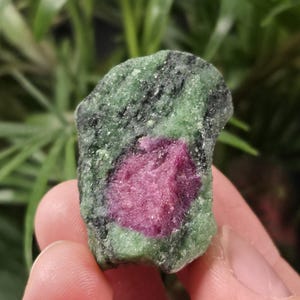 May include: A rough-cut ruby in zoisite gemstone. The stone features a vibrant pink ruby center surrounded by a green and black zoisite matrix. The natural stone is held in a hand, with a blurred green background.