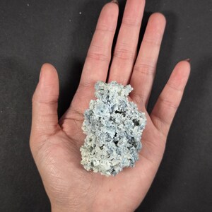 May include: A light blue and white mineral specimen with a porous, textured surface. The mineral is held in a hand against a black background. The mineral appears to be a cluster of small, interconnected formations.