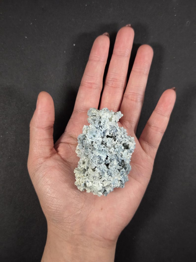 May include: A light blue and white mineral specimen with a porous, textured surface. The mineral is held in a hand against a black background. The mineral appears to be a cluster of small, interconnected formations.