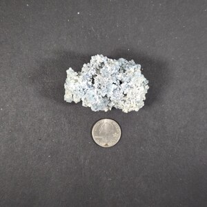 May include: A light blue, porous mineral specimen with a quarter coin for size comparison. The mineral has a rough, textured surface and a clustered formation. The background is a dark gray surface.