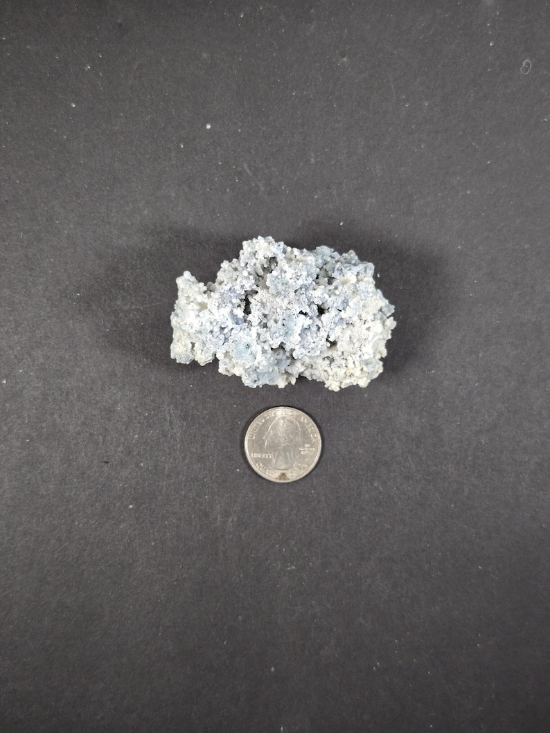 May include: A light blue, porous mineral specimen with a quarter coin for size comparison. The mineral has a rough, textured surface and a clustered formation. The background is a dark gray surface.