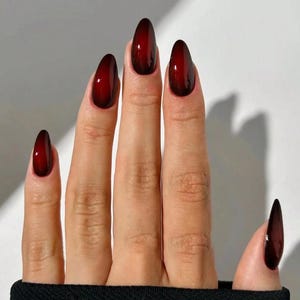 May include: A hand with almond-shaped nails featuring a glossy, deep oxblood red polish with a darker, near-black gradient effect at the center of each nail. The fingers are positioned against a plain background, showcasing the smooth, reflective finish.