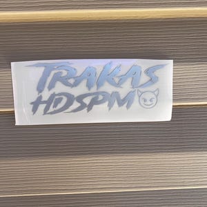 Może przedstawiać: Biała naklejka z tekstem "TRAKAS HDSPM" w stylizowanym, szarym foncie. Mała, szara diabelska twarz z uśmiechem znajduje się po prawej stronie tekstu.