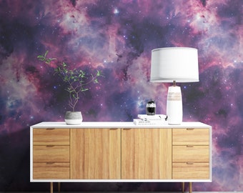 May include: A white dresser with three drawers and wooden fronts sits in front of a wall with a purple and pink galaxy print wallpaper. A small potted plant and a white lamp with a white lampshade are on top of the dresser.