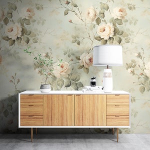 May include: A white dresser with three drawers and wooden fronts sits in front of a wall with a floral wallpaper featuring white roses and green leaves.