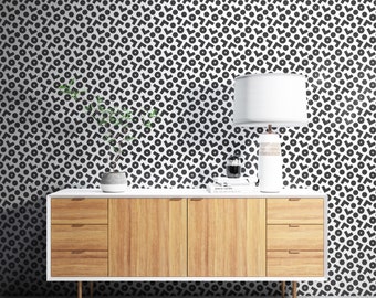 May include: A mid-century modern style dresser with three drawers and white legs. The dresser is in front of a white wall with a black and white geometric pattern wallpaper. A lamp with a white shade and a potted plant are on top of the dresser.