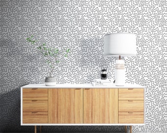 May include: A white dresser with three drawers and a white top. The dresser has a wood grain finish and brass handles. The dresser is in front of a white wall with a black and white geometric pattern.
