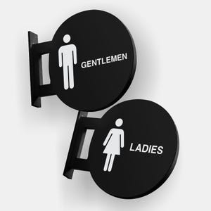 May include: Two black circular signs with white stick figures and text. The sign on the left says "GENTLEMEN" and the sign on the right says "LADIES".