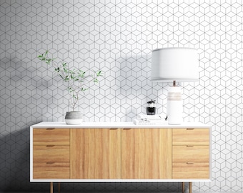 May include: A white dresser with three drawers and a white top. The dresser has a wood grain finish and brass handles. A white lamp with a white shade sits on the dresser, along with a small plant in a white pot and a stack of books. The dresser is in front of a white wall with a geometric pattern.