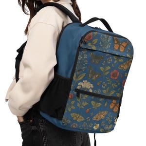 May include: A blue backpack with a floral and butterfly pattern. The backpack has a black side pocket and a black handle. The floral pattern includes orange, white, and yellow flowers. The backpack is being worn by a person.