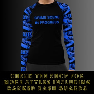 May include: Black rash guard with blue caution tape design on the sleeves. The shirt has the words "CRIME SCENE IN PROGRESS" in blue on the front. The bottom of the image has text that says "CHECK THE SHOP FOR MORE STYLES INCLUDING RANKED RASH GUARDS."