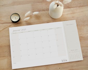 May include: A beige 2025 calendar showing January, with a small February calendar. A small candle and a white vase with dried flowers are on a wooden surface.