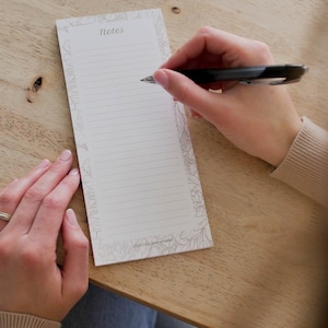 Peut inclure: Un bloc-notes rectangulaire avec le mot "Notes" en haut, avec du papier ligné et une bordure florale. Un stylo noir est tenu au-dessus du bloc-notes, prêt à écrire. Le bloc-notes repose sur une surface en bois clair.