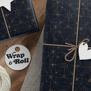 Puede incluir: Regalos envueltos con papel azul marino y un patrón geométrico dorado. Cada paquete está atado con hilo y una etiqueta blanca. También se ven un ovillo de hilo, tijeras y etiquetas. La etiqueta redonda dice "Wrap & Roll".