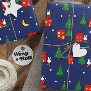 May include: Gift-wrapped presents with navy blue paper featuring a repeating pattern of houses, Christmas trees, and a bunny. The gifts are tied with twine and adorned with star and heart-shaped tags. A spool of twine, scissors, and tags are also visible.