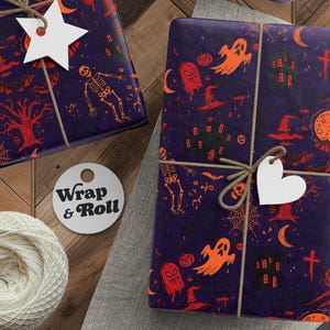 A gift wrapped in spooky Halloween wrapping paper. Features orange and red illustrations of skeletons, ghosts, haunted houses, and skulls on a dark purple background. The present is tied with twine and surrounded by festive pine branches.
