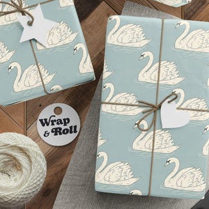 Puede incluir: Papel de regalo azul claro con un patrón repetitivo de cisnes blancos nadando en el agua. Los paquetes están atados con cordel marrón e incluyen etiquetas de regalo en forma de estrella y corazón. Hay un ovillo de cordel y unas tijeras negras.