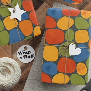 Puede incluir: Regalos envueltos con patrones abstractos de guijarros coloridos en naranja, amarillo, verde y azul sobre un fondo gris. Cada paquete está atado con hilo y una etiqueta blanca. También se ven un carrete de hilo, tijeras y etiquetas.