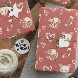 Puede incluir: Regalos envueltos con un patrón repetido de mapaches, corazones, calcetines y caras de oso sobre papel rojo. Los regalos están atados con hilo y adornados con etiquetas blancas en forma de corazón. También se ven un carrete de hilo, unas tijeras y una etiqueta que dice "Wrap & Roll".