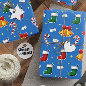 Könnte beinhalten: Verpackte Geschenke mit blauem Geschenkpapier mit Weihnachtssocken, Glocken, Zuckerstangen und Sternen. Die Geschenke sind mit Bindfaden gebunden und mit weißen Stern- und herzförmigen Anhängern versehen. Eine Garnrolle, eine Schere und ein "Wrap & Roll"-Etikett sind ebenfalls vorhanden.