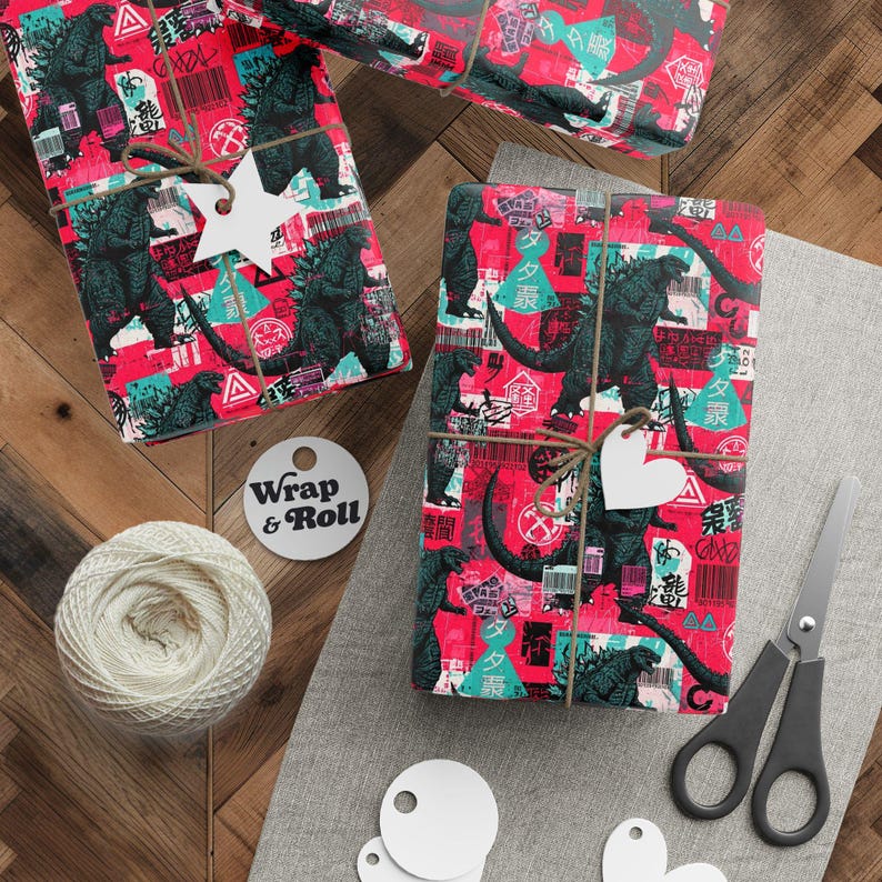 A gift wrapped in street art style Godzilla wrapping paper, featuring a chaotic collage of the monster, graffiti, Japanese text, and warning labels in a bold red, teal, and black color scheme. The present is tied with twine and surrounded by pine branches.