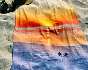 May include: A beach towel featuring a sunset and ocean scene. The towel is folded and lying on a sandy beach. A brown hat and sandals are lying on the sand next to the towel.
