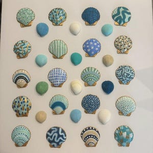 May include: A collection of painted seashells in various colors and patterns, including blue, green, and gold, arranged on a white background. The seashells are of different sizes and shapes, creating a decorative display.