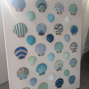 May include: A white rectangular canvas featuring a collection of individual seashells arranged in a grid. Each shell is painted in various shades of blue, teal, and white, showcasing unique patterns including stripes, dots, and floral motifs.