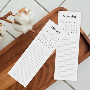 May include: Two vertical white paper calendar bookmarks featuring black text. One displays the month of September and the other October, each with a grid of dates and a series of horizontal lines below for notes, arranged on a wooden tray.