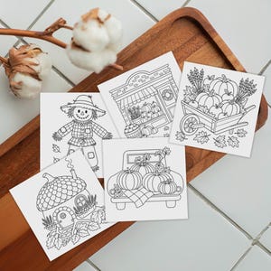 May include: Five square white paper cards featuring black line art illustrations of autumn themes including a scarecrow, a storefront with pumpkins, a wheelbarrow filled with produce, a truck carrying pumpkins, and an acorn-shaped house.