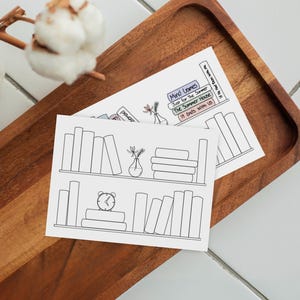 May include: Two white rectangular cards featuring black line art illustrations of bookshelves. One card shows books, a vase with flowers, and an alarm clock. The other card displays a stack of books with titles including Mind Games and It Ends With Us.