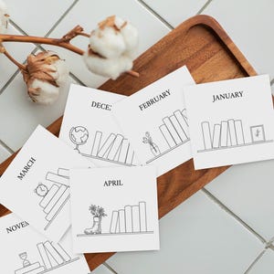May include: A set of square white cards featuring black line art illustrations of bookshelves labelled with months including January, February, March, April, and December. Each card displays a unique shelf arrangement with books and decorative items.