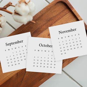 May include: Three square white paper calendar cards for September, October, and November with black serif text and numbers. The cards are arranged on a brown wooden tray next to a dried cotton stem with white fluffy bolls on a white tiled surface.