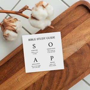 May include: A square white card featuring the black text BIBLE STUDY GUIDE and the acronym SOAP with definitions for Scripture, Observation, Application, and Prayer. The card is positioned near a brown wooden tray and white cotton stems.