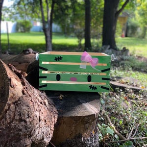 May include: A green wooden crate with a painted monster face design. The crate features black accents, pink cheeks, and a pink bow. The crate is sitting on a tree stump outdoors.