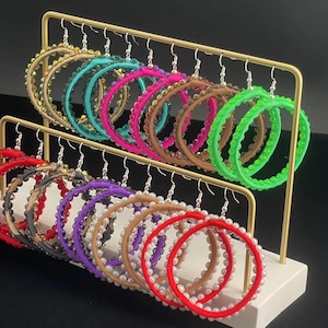 Może przedstawiać: Prezentacja kolczyków koła w różnych kolorach, w tym czerwonym, fioletowym, brązowym i zielonym. Każdy kolczyk jest ozdobiony małymi, białymi koralikami. Kolczyki są ułożone na złotym metalowym stojaku.