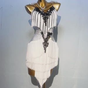 Puede incluir: Vestido de baile blanco con detalles de encaje y flecos negros. El vestido tiene un escote en V y un corpiño ajustado. La falda está hecha de capas de flecos y es asimétrica.
