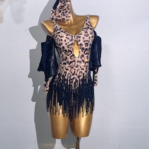 Op de afbeelding: Een danskostuum met luipaardprint met een diepe halslijn en zwarte franjes. Het kostuum bevat een bijpassende hoofdbekleding en off-the-shoulder mouwen. De mannequin is goudkleurig en de achtergrond is lichtgrijs.