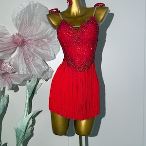 Könnte beinhalten: Ein rotes Tanzkleid mit einem bestickten Oberteil und einem Fransenrock wird auf einer goldenen Schaufensterpuppe ausgestellt. Das Kleid hat dünne Träger und passende Perlenakzente an den Schultern. Ein passender Kopfschmuck befindet sich ebenfalls auf der Schaufensterpuppe.